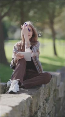 Öğrenci, parkta alçak bir duvarda otururken notlar yazıyor.