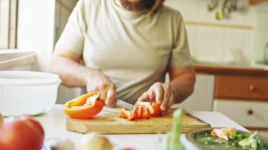 Adam, mutfak ortamında ahşap bir kesme tahtasının üzerinde portakal dolmalık biberi kesiyor.