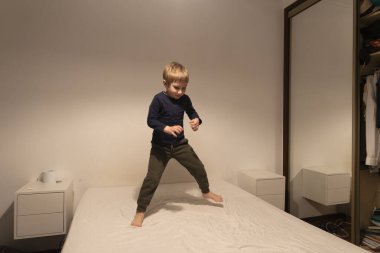 Sarışın çocuk modern yatak odasında gündelik kıyafetler giyerek yatakta zıplarken eğleniyor.