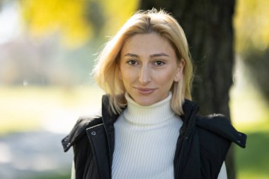 Balıkçı yaka kazak ve siyah yelek giyen genç bir kadın güneşli bir sonbahar gününde parkta gülümsüyor.