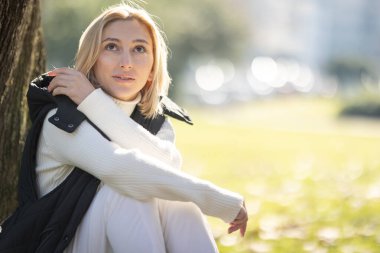 Siyah yelek ve beyaz balıkçı yaka kazak giyen genç sarışın kadın sonbahar günü parkta ağacın yanında oturuyor.