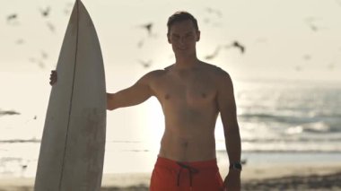 Gün batımında sörf tahtasıyla sahilde duran genç sörfçü.