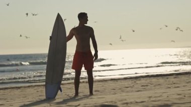 Gün batımında sörf tahtasıyla sahilde duran genç sörfçü okyanusa bakıyor.