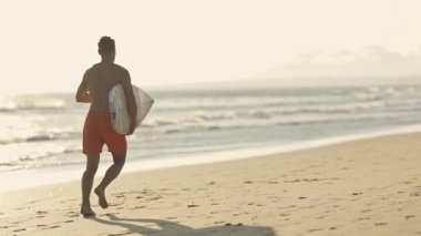 Gün doğumunda sörf tahtasıyla sahilde koşan genç sörfçü.