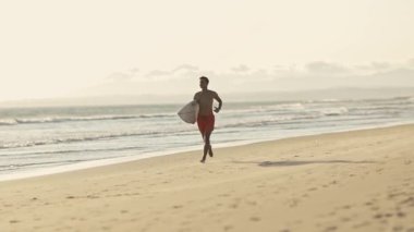 Gün batımında sörf tahtasıyla sahilde koşan genç sörfçü.