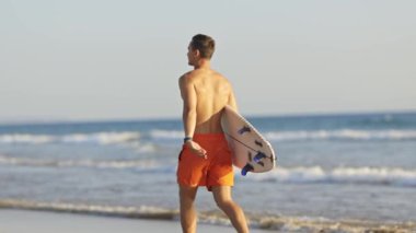 Genç sörfçü elinde sörf tahtasıyla sahilden uzaklaşıyor.