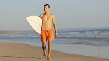 Genç sörfçü sörf tahtasıyla sahilde yürüyor.