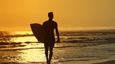 Gün batımında okyanustan çıkan bir sörfçünün silüeti.
