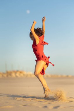 Gün batımında kumlu bir plajda zarif bir atlayış yapan kadın dansçı kırmızı bir elbise giyiyor.