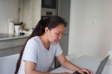 Laptop kullanan, ev mutfağından uzaktan çalışan odaklanmış genç Asyalı kadın.