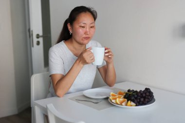 Genç Asyalı kadın evdeki masada taze meyve tabağıyla kahve içiyor.
