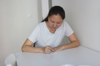 Genç Asyalı bir kadın not defterine kalemle notlar yazıyor. Evdeki beyaz bir masada oturuyor.
