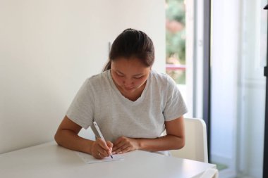 Asyalı bir kadın beyaz bir masada otururken kağıt ve kalemle not alıyor.