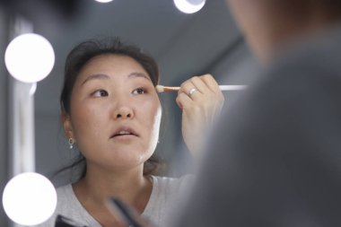 Genç Asyalı bir kadın yüzüne makyaj yapıyor. Işıklı bir aynanın önünde fırça kullanıyor.