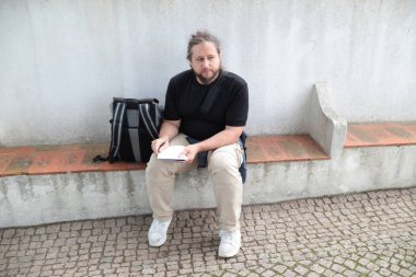 Erkek turist, yanında sırt çantasıyla bankta otururken deftere notlar yazıyor.