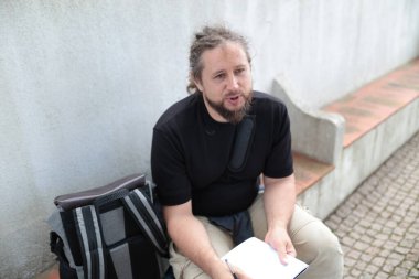 Erkek üniversite öğrencisi dışarıda ders çalışıyor ve konuşuyor. Sırt çantası ve not defteriyle merdivenlerde oturuyor.