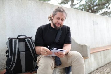 Sakallı adam şehir merkezindeki basamaklarda otururken deftere notlar yazıyor.