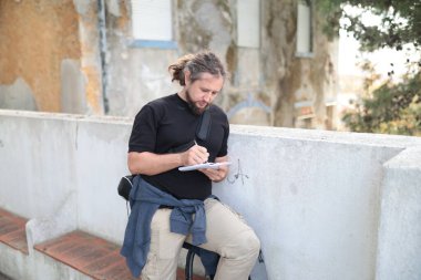 Erkek gazeteci dışarıda deftere notlar alıyor, kentsel ortamda işine odaklanıyor.