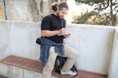 Erkek gazeteci şehir ortamında alçak bir duvara yaslanırken not defterine notlar yazıyor.