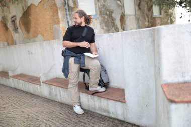 Erkek gazeteci açık hava bankında otururken not alıyor.