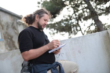 Gazeteci açık havada notlar alıyor, bir not defterine yazıyor, güncel bir olayı gözlemliyor ve rapor ediyor