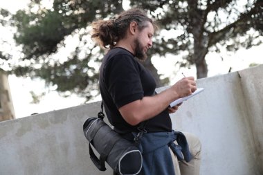 Arazi mühendisi dışarıda çalışırken bir not defterine notlar yazıyor. Kalem kullanıyor ve tripod çanta taşıyor.