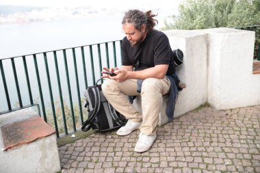 Akıllı telefon kullanan erkek turist, arka planda deniz manzarası ile dinlenirken