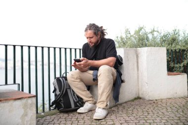 Akıllı telefon kullanan erkek turist yanında sırt çantasıyla manzaraya bakıyor.