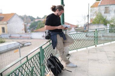 Küçük bir Avrupa kasabasına bakan balkonda akıllı telefon kullanan ve harita okuyan genç bir turist.