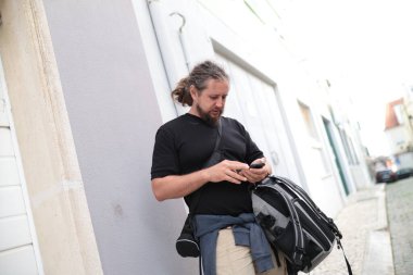 Akıllı telefon kullanan ve şehir sokaklarında yürürken sırt çantası taşıyan bir erkek turist.