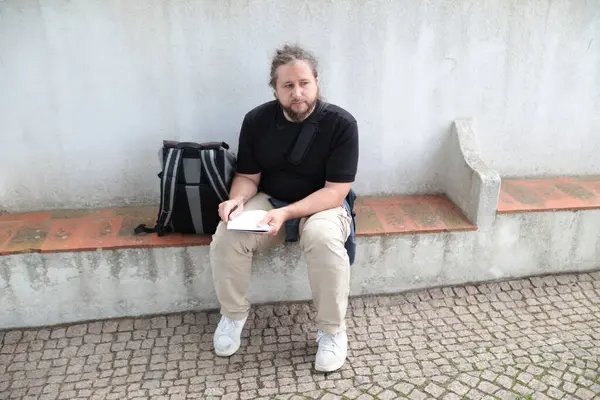 Erkek turist, yanında sırt çantasıyla bankta otururken deftere notlar yazıyor.