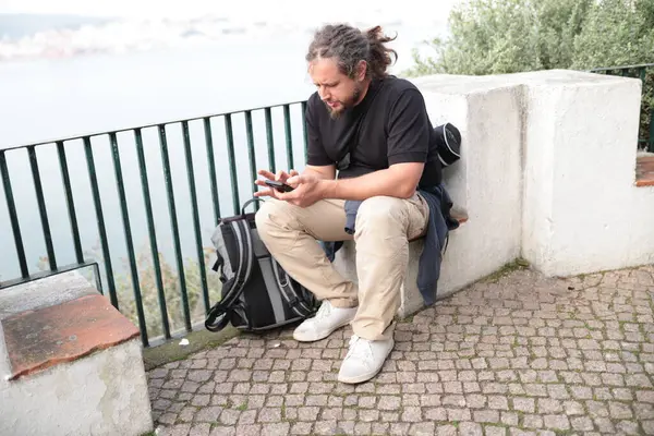Akıllı telefon kullanan erkek turist, arka planda deniz manzarası ile dinlenirken