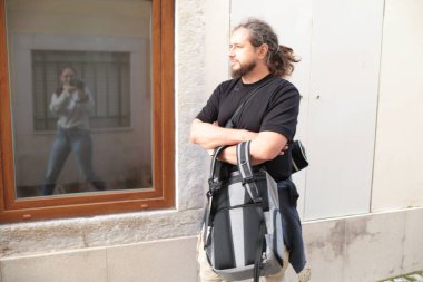 Kolları çapraz duran, sırt çantalı ve kamera çantalı bir erkek fotoğrafçı. Yansıması yakın bir pencerede görülüyor.