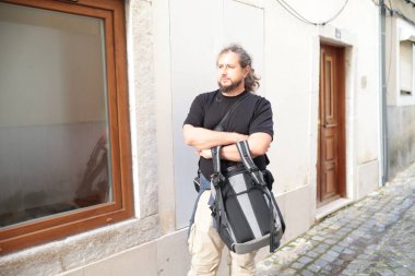 Kolları çapraz bekleyen, siyah gömlekli, bej pantolonlu, sırt çantalı, kaldırım taşlı bir sokakta bekleyen bir erkek fotoğrafçı.