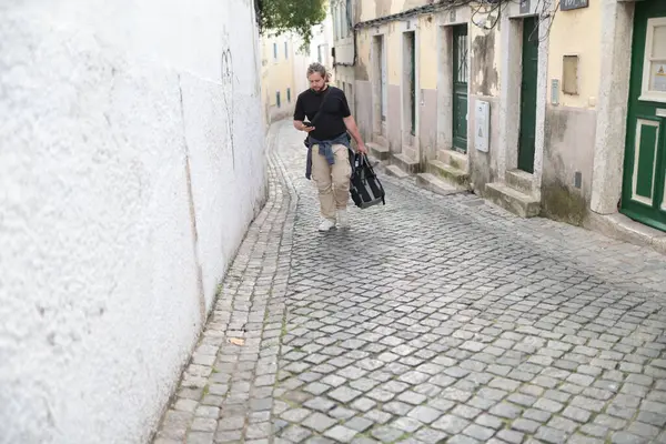 Lizbon, Portekiz 'de dar bir kaldırım taşı caddesinde yürürken akıllı telefon kullanan erkek turist