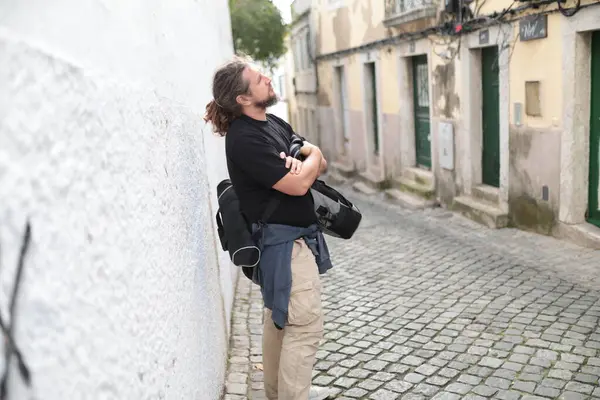 Fotoğraf makinesi çantası ve kolları çaprazlanmış Lizbon, Portekiz 'de dar bir kaldırım taşı sokakta beyaz bir duvara yaslanmış.