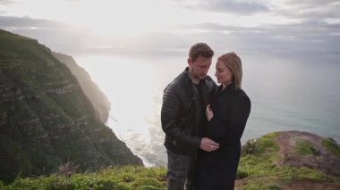 Gün batımında okyanusa bakan bir uçurumda öpüşen romantik çift.
