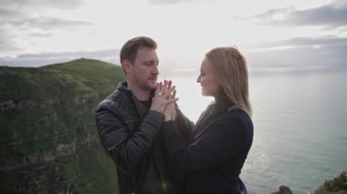 Sevgili çift el ele tutuşup gün batımında okyanusa bakan bir uçurumda birbirlerine bakıyorlar.