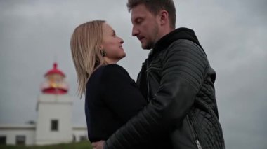 Çift, bulutlu bir gökyüzünün altında, resim gibi bir deniz fenerinin yanında samimi bir kucaklaşmayı paylaşıyor, romantizm ve dinginliği aktarıyor.