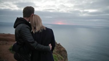 Genç çift gün batımında okyanusa bakan bir uçurumda romantik bir anın tadını çıkarıyor.