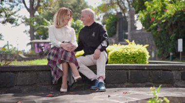 Alçak bir duvarda oturan olgun bir çift, güneşli madeira botanik bahçesinde mutlu bir şekilde sohbet ediyorlar.