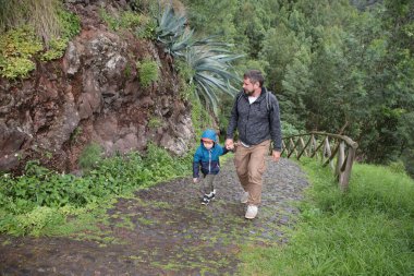 Baba ve oğul, bereketli bir ormanın canlı yeşillikleriyle çevrili pitoresk bir kaldırım yolu boyunca sakin bir yürüyüş yapıyorlar.