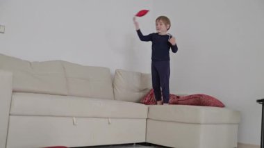 Sarışın bir çocuk kırmızı yıldız şeklinde bir balonla beyaz bir kanepede zıplayıp oynuyor.