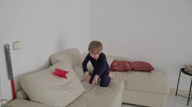 Genç çocuk eğleniyor, zıplıyor ve parlak bir oturma odasında rahat bir kanepede oynuyor.