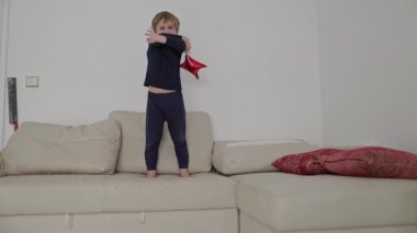 Sarışın çocuk beyaz bir kanepede dururken kırmızı yıldız şekilli balonlarla oynuyor.