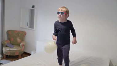 Güneş gözlüğü takan mutlu çocuk yatağa atlıyor ve beyaz balonlarla oynuyor.