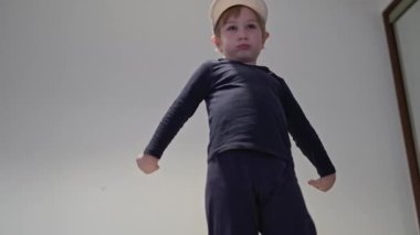 Beyaz şapkalı ve koyu renk elbiseli bir çocuk yatak odasında dans figürleri yapıyor.