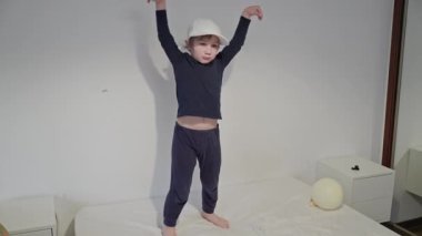 Enerjik çocuk yatak odasındaki yatakta dans figürleri yapıyor, beyzbol şapkası takıyor.