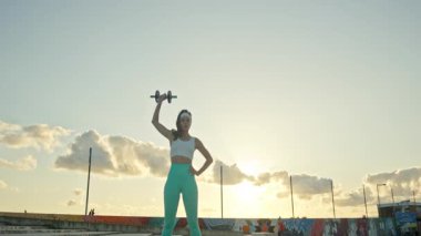 Atletik kadın gün batımı egzersizi sırasında bir halterle kol egzersizi yapıyor.