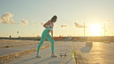 Spor salonundaki kadın, gün batımında kaldırım kenarında halterlerle egzersiz yapıyor.